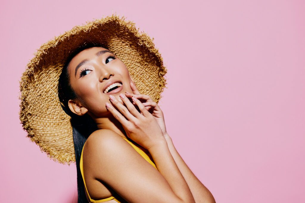 Donna sorridente con cappello di paglia su sfondo rosa, espressione luminosa ed estiva – preparazione all’estate 2025, bellezza viso e skincare.