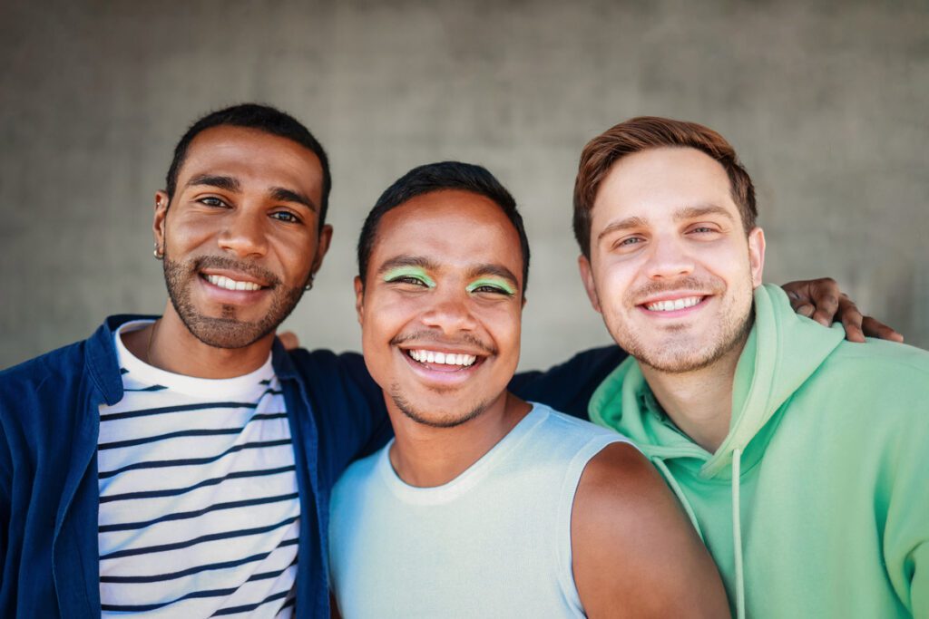 Tre giovani uomini sorridenti si abbracciano con naturalezza e complicità. Espressione di inclusività, identità e libertà estetica. By Centro Medico Chirurgico Matteotti.